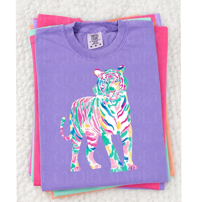 May include: A lilac Comfort Colors t-shirt with a colourful tiger illustration. The tiger is rendered in bright pink, green, blue, and yellow. The shirt is stacked with other brightly coloured shirts.