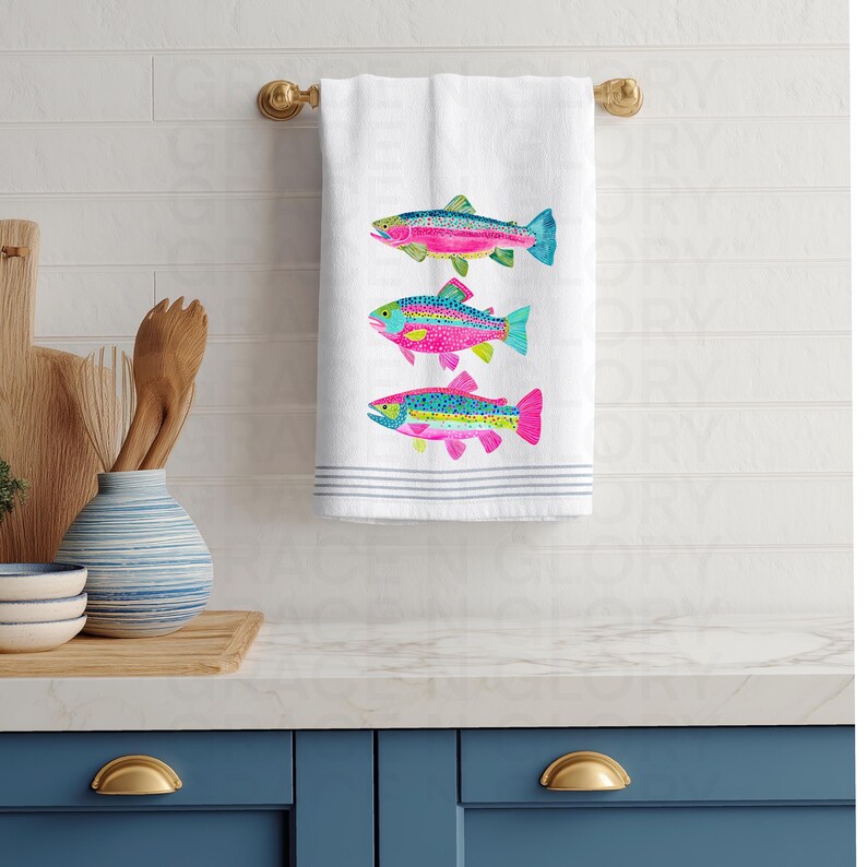 May include: White hand towel with three embroidered rainbow trout. The fish have pink, blue, green, and yellow details. The towel has blue stripes at the bottom and is hanging on a gold towel rack.