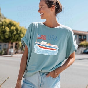 Op de afbeelding: Lichtblauw T-shirt met een illustratie van een boot, een Amerikaanse vlag en het woord "USA". De boot heeft rode, witte en blauwe strepen. Het T-shirt heeft opgerolde mouwen. Het T-shirt is gemaakt van zacht materiaal.