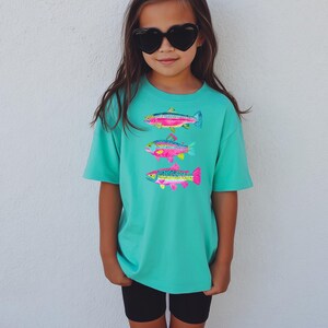 May include: A turquoise t-shirt featuring three colorful fish designs. The fish are in shades of pink, blue, yellow, and green. The shirt is paired with black shorts and heart-shaped sunglasses.
