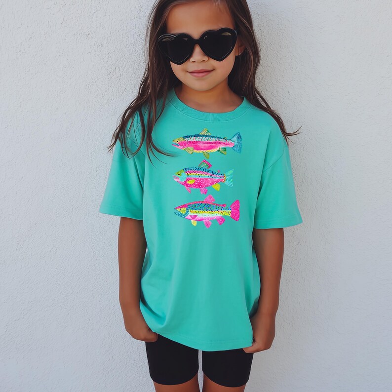 May include: A turquoise t-shirt featuring three colorful fish designs. The fish are in shades of pink, blue, yellow, and green. The shirt is paired with black shorts and heart-shaped sunglasses.