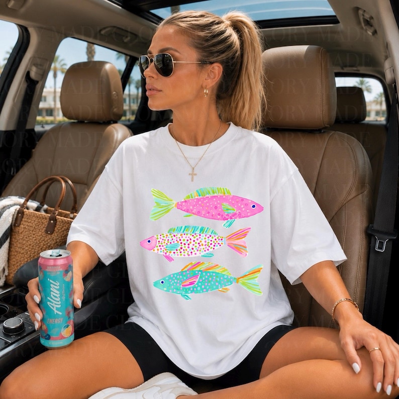 May include: White t-shirt featuring three colorful fish designs. The fish are pink, blue, and a mix of colors, with patterned scales and bright fins. The shirt is a casual, oversized style.