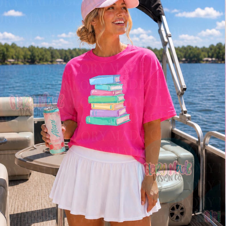May include: A bright pink t-shirt featuring a stack of colorful books. The books are in shades of blue, pink, green, yellow, and purple. The shirt is paired with a white tennis skirt. The image is taken outdoors near a lake.