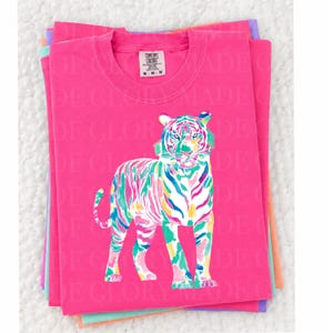 May include: A stack of pink Comfort Colors t-shirts with a colourful tiger design. The tiger is depicted with vibrant strokes of blue, green, yellow, and pink. The shirts have a crew neck and the Comfort Colors tag is visible.