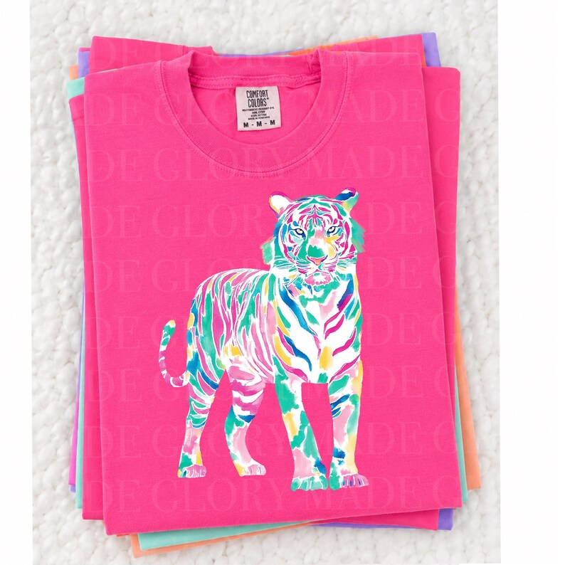 May include: A stack of pink Comfort Colors t-shirts with a colourful tiger design. The tiger is depicted with vibrant strokes of blue, green, yellow, and pink. The shirts have a crew neck and the Comfort Colors tag is visible.
