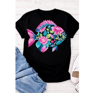 May include: Black t-shirt featuring a colorful fish design. The fish is adorned with pink flowers, teal and blue accents, and green leaves. The fish's tail and fins are pink. The shirt is paired with blue jeans and white sneakers.
