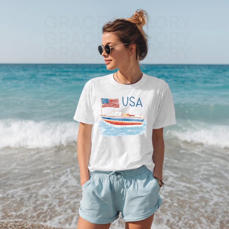 Op de afbeelding: Wit T-shirt met een aquarel illustratie van een boot, een Amerikaanse vlag en het woord "USA" in blauw. De persoon draagt het T-shirt op het strand, gecombineerd met een blauwe short en zonnebril.