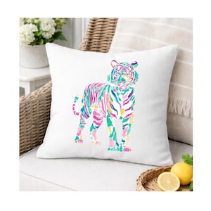 May include: A white square cushion with a vibrant watercolour tiger design. The tiger is rendered in pink, green, blue, and yellow hues. The cushion is set on a white seat, with a wicker chair and decorative elements visible in the background.