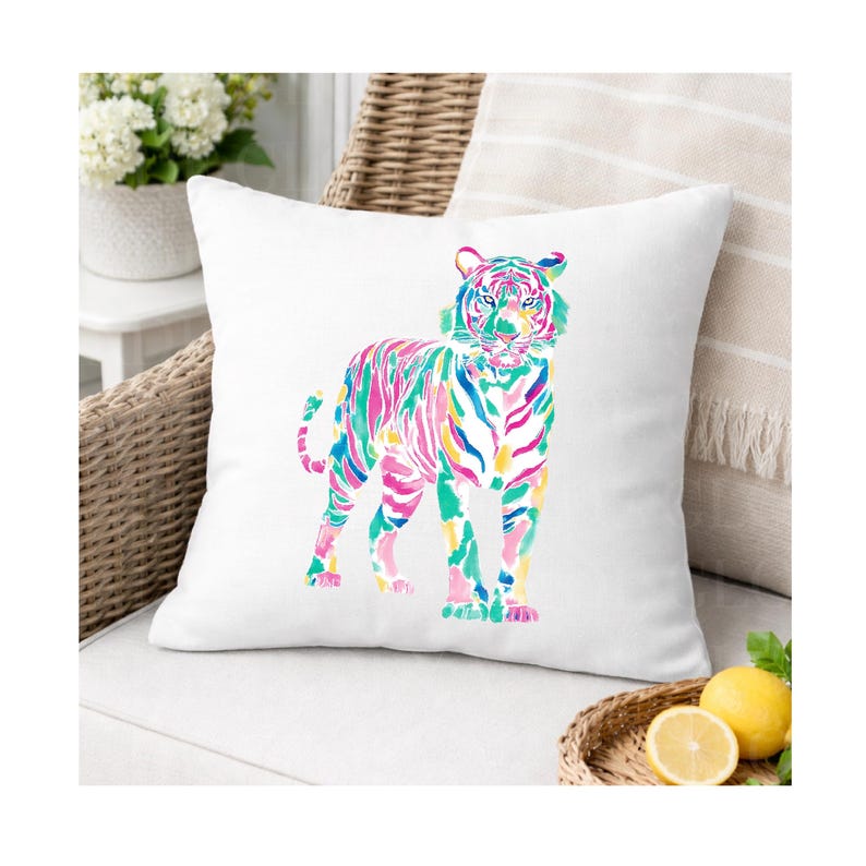 May include: A white square cushion with a vibrant watercolour tiger design. The tiger is rendered in pink, green, blue, and yellow hues. The cushion is set on a white seat, with a wicker chair and decorative elements visible in the background.