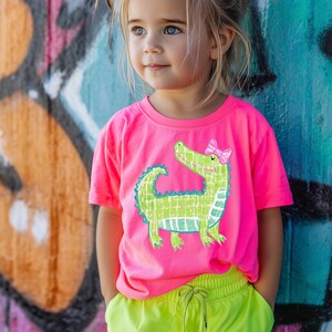 May include: A young child wearing a bright pink t-shirt with a green alligator graphic and a pink bow. The child is also wearing neon yellow shorts. The background features graffiti art.