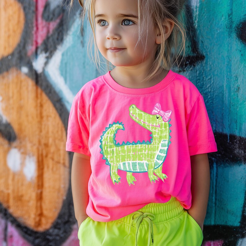May include: A young child wearing a bright pink t-shirt with a green alligator graphic and a pink bow. The child is also wearing neon yellow shorts. The background features graffiti art.