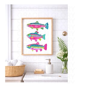 May include: A framed watercolor print featuring three colorful fish. Each fish has a unique pattern of pink, blue, green, and yellow, set against a white background. The artwork is displayed in a light brown wooden frame, suitable for bathroom or coastal decor.