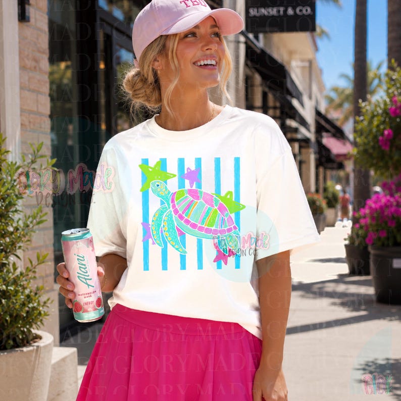 May include: A white short-sleeve t-shirt featuring a graphic of a sea turtle in shades of pink, green, and blue, set against vertical blue stripes and small star shapes. The shirt is paired with a light pink baseball cap and a bright pink skirt.