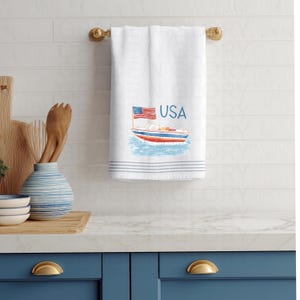 Op de afbeelding: Witte katoenen handdoek met een nautisch ontwerp met een boot, de Amerikaanse vlag en het woord "USA" in blauw. De handdoek heeft blauwe strepen aan de onderkant en hangt aan een gouden handdoekrek.