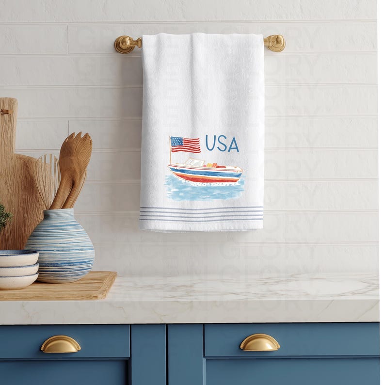 Op de afbeelding: Witte katoenen handdoek met een nautisch ontwerp met een boot, de Amerikaanse vlag en het woord "USA" in blauw. De handdoek heeft blauwe strepen aan de onderkant en hangt aan een gouden handdoekrek.