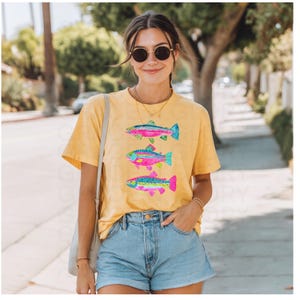 May include: Yellow t-shirt featuring three colorful fish designs. Each fish has a unique pattern with shades of pink, blue, and green. The shirt is a casual style, perfect for everyday wear.