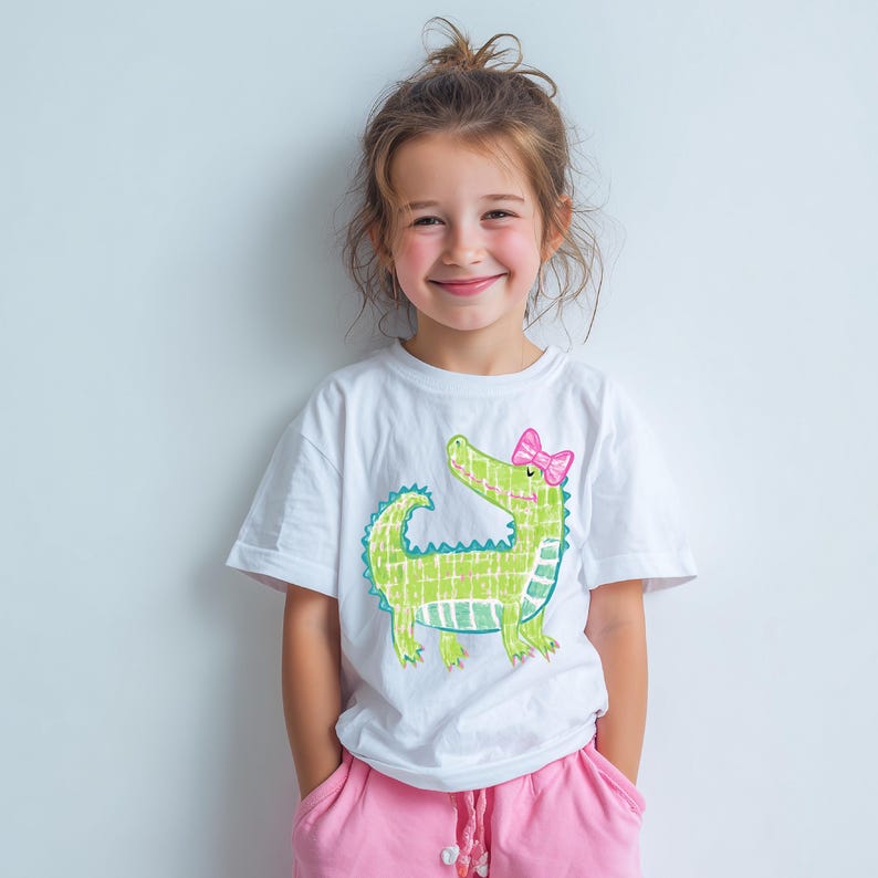 May include: A white t-shirt featuring a green alligator illustration with a pink bow. The alligator design is outlined in blue and has a grid-like pattern. The shirt is paired with pink shorts.