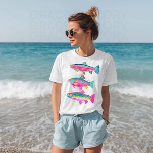 May include: White t-shirt featuring three colorful fish illustrations. The fish have pink, blue, green, and yellow accents. The shirt is worn by a person on a beach with blue water and sky in the background.