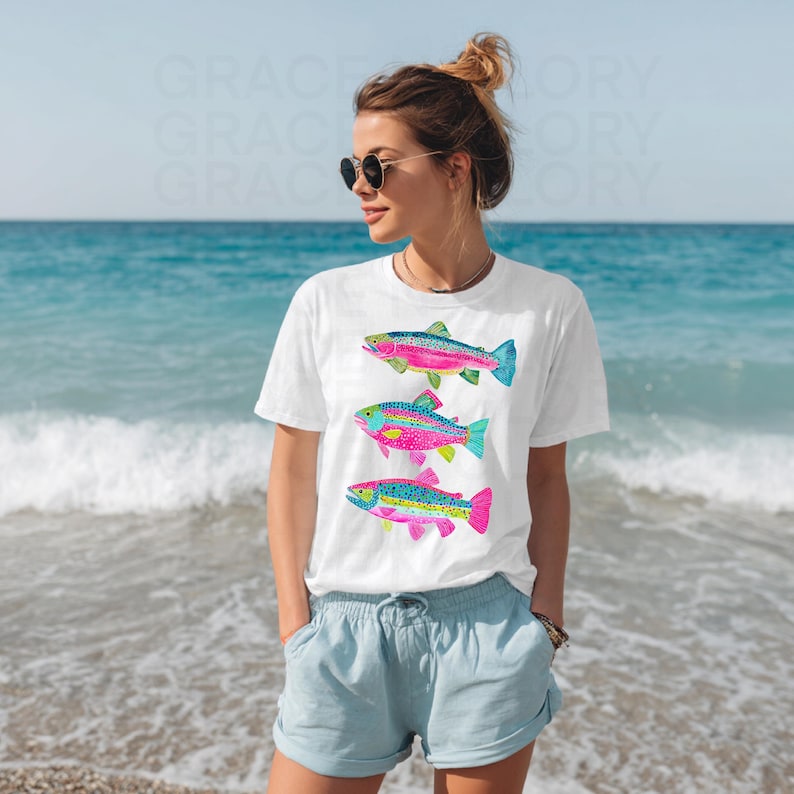 May include: White t-shirt featuring three colorful fish illustrations. The fish have pink, blue, green, and yellow accents. The shirt is worn by a person on a beach with blue water and sky in the background.