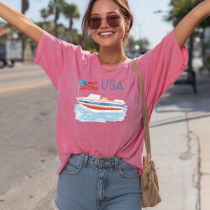 Op de afbeelding: Roze t-shirt met een afbeelding van een boot, de Amerikaanse vlag en het woord "USA". De boot is afgebeeld in blauw, rood en wit. Het shirt wordt gecombineerd met een blauwe spijkerbroek.