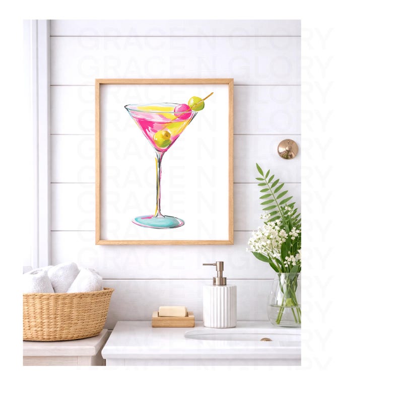 May include: A framed watercolor print of a martini cocktail. The drink is pink and yellow, garnished with green olives and a pink cherry. The artwork is in a light wood frame, displayed in a bathroom setting with a white sink and accessories.