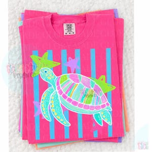 May include: A bright pink short-sleeve t-shirt featuring a graphic of a sea turtle with a multi-colored striped shell. The design includes vertical light blue stripes and several lime green and purple stars. The neck label reads Comfort Colors M.