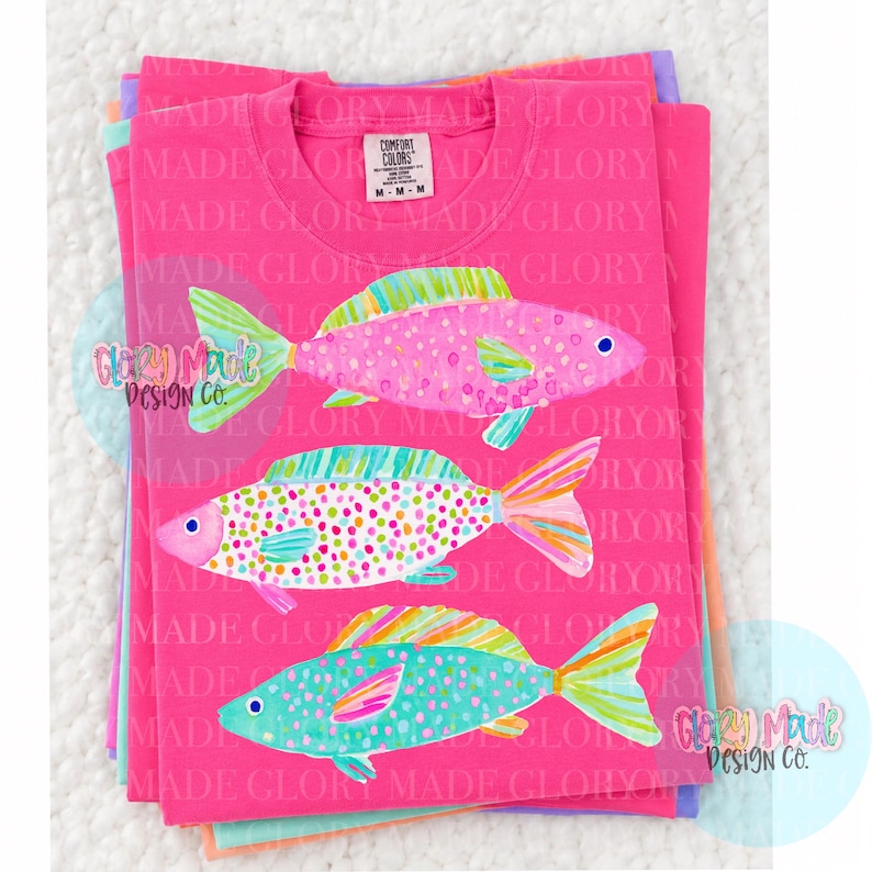 May include: A stack of pink t-shirts with a colorful fish design. Each fish is a different color and pattern, with details in pink, green, blue, and yellow. The shirts have the text "MADE GLORY" and "COMFORT COLORS" on them.