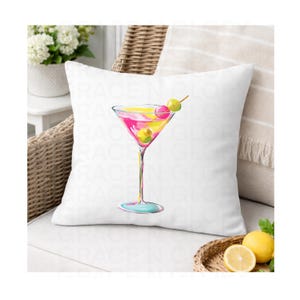 May include: White square throw pillow featuring a watercolor illustration of a pink and yellow martini with green olives. The cocktail glass has a long stem and a light blue base. The pillow is on a wicker chair.
