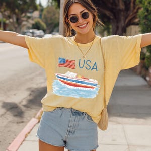 Op de afbeelding: Geel T-shirt met een afbeelding van een boot, de Amerikaanse vlag en het woord "USA". Het shirt wordt gecombineerd met een spijkerbroek. De foto is buiten op een zonnige dag gemaakt.