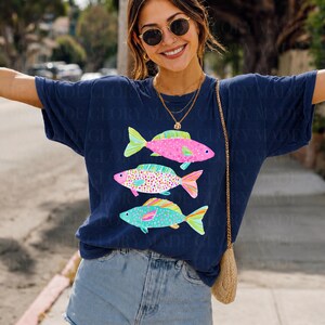 May include: Navy blue t-shirt featuring three colorful fish illustrations. The fish are pink, green, and blue with patterned details. The shirt has a relaxed fit and is paired with denim shorts.