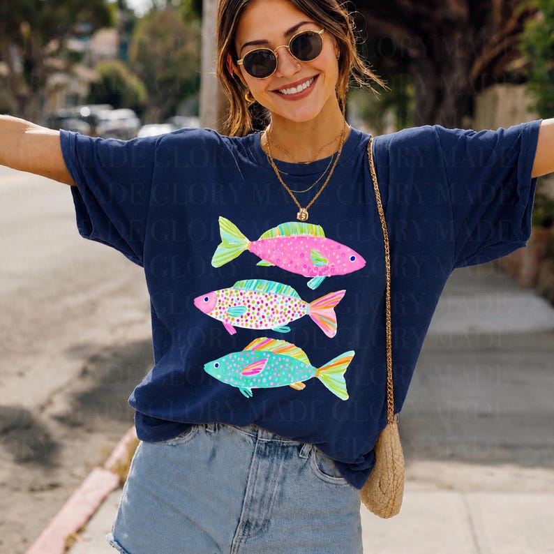 May include: Navy blue t-shirt featuring three colorful fish illustrations. The fish are pink, green, and blue with patterned details. The shirt has a relaxed fit and is paired with denim shorts.