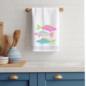May include: White hand towel with three colorful fish designs. The fish are pink, turquoise, and a speckled pattern, with green and orange accents. The towel has gray stripes at the bottom and is hanging on a gold towel bar.