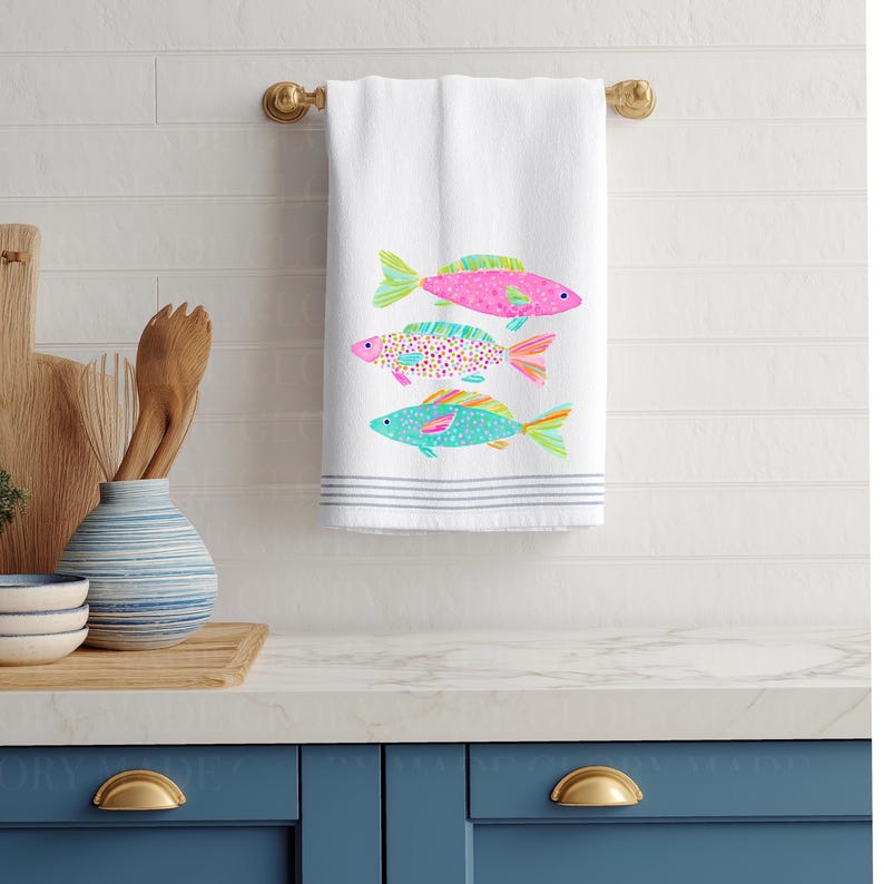 May include: White hand towel with three colorful fish designs. The fish are pink, turquoise, and a speckled pattern, with green and orange accents. The towel has gray stripes at the bottom and is hanging on a gold towel bar.