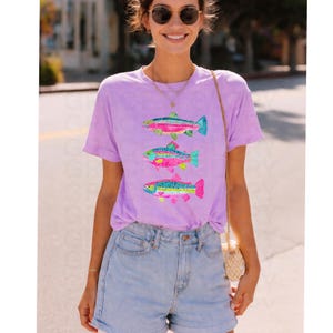 May include: A light purple t-shirt featuring three colorful fish designs. Each fish has a unique pattern with shades of pink, blue, yellow, and green. The shirt is paired with denim shorts.