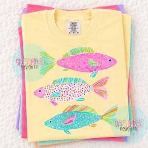 May include: Yellow Comfort Colors t-shirt featuring three colorful fish illustrations. The fish are pink, teal, and multi-colored with dot and scale patterns. The shirt is folded with other pastel colored shirts.