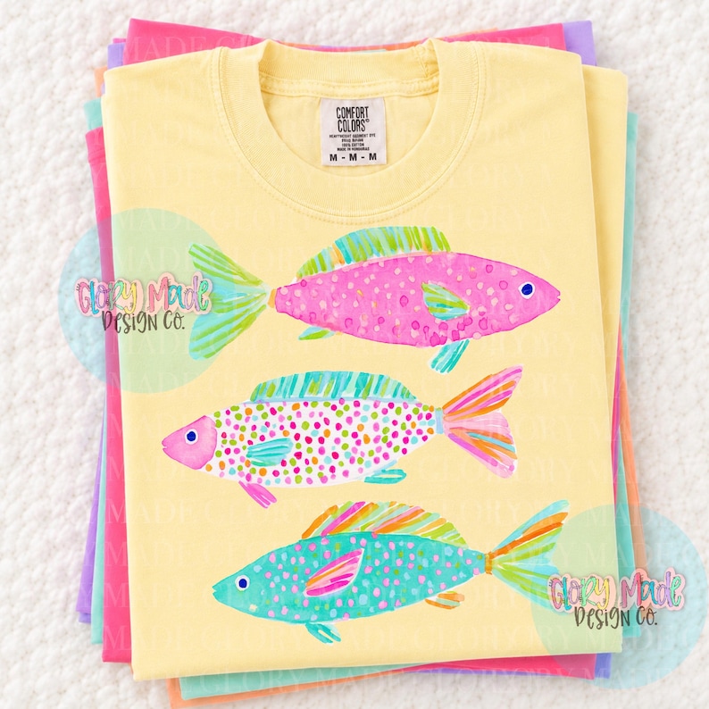 May include: Yellow Comfort Colors t-shirt featuring three colorful fish illustrations. The fish are pink, teal, and multi-colored with dot and scale patterns. The shirt is folded with other pastel colored shirts.