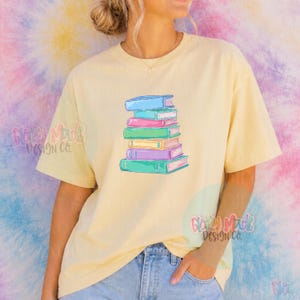 May include: A light yellow t-shirt featuring a stack of colorful books. The books are in shades of blue, pink, green, and purple. The shirt is paired with blue jeans. The design is a whimsical illustration of a book lover's dream.