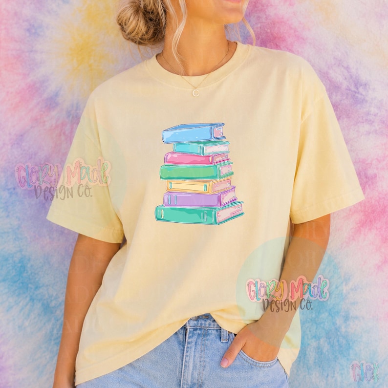 May include: A light yellow t-shirt featuring a stack of colorful books. The books are in shades of blue, pink, green, and purple. The shirt is paired with blue jeans. The design is a whimsical illustration of a book lover's dream.