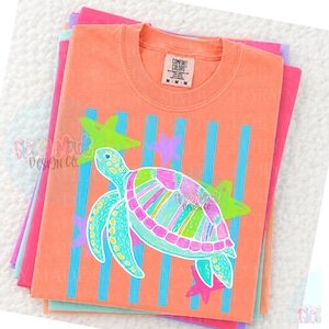 May include: A peach-colored crew neck t-shirt with a Comfort Colors label at the collar. The graphic features a multi-colored sea turtle in shades of pink, green, and blue, set against a background of vertical blue stripes and several star shapes.