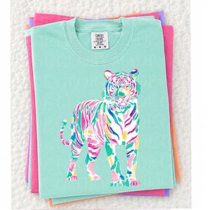 May include: A light teal Comfort Colors t-shirt with a colourful tiger illustration. The tiger is depicted with vibrant pink, green, blue, and yellow stripes. The shirt is stacked on top of other folded shirts in shades of pink and purple.