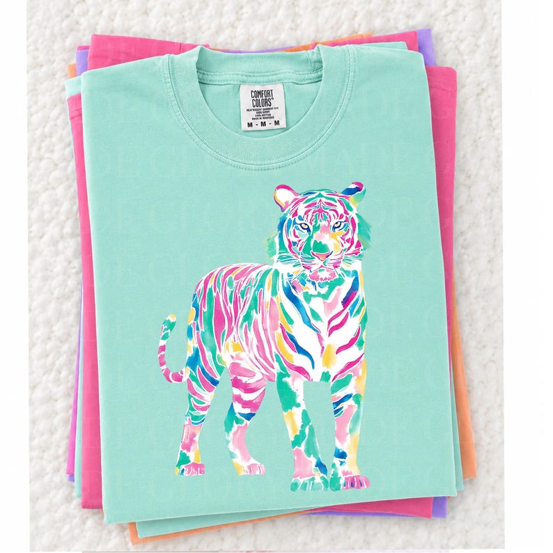 May include: A light teal Comfort Colors t-shirt with a colourful tiger illustration. The tiger is depicted with vibrant pink, green, blue, and yellow stripes. The shirt is stacked on top of other folded shirts in shades of pink and purple.