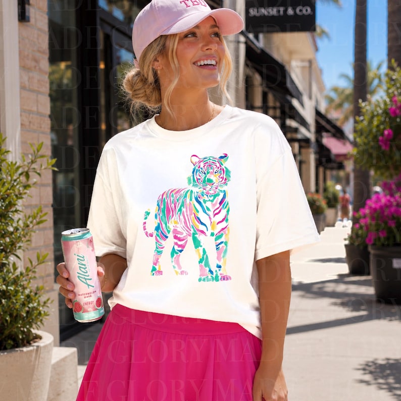 May include: Cream-coloured t-shirt with a colourful tiger illustration. The tiger is rendered in strokes of pink, blue, green, and yellow. The person is wearing a pink baseball cap and a pink skirt. A can of Alani watermelon drink is held in the hand.