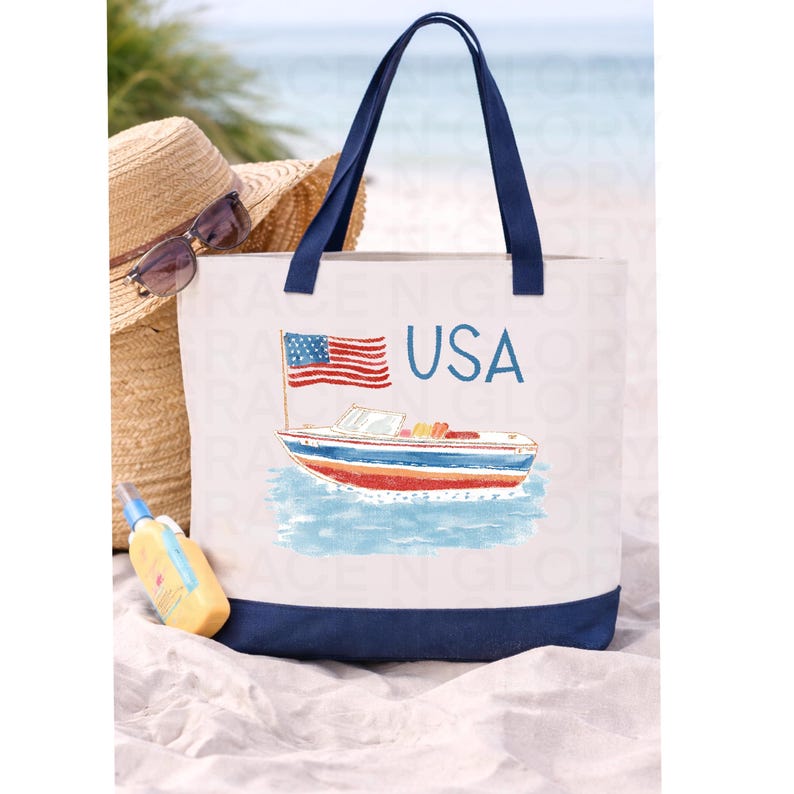 Op de afbeelding: Witte canvas draagtas met marineblauwe handvatten en basis. De tas heeft een kleurrijke illustratie van een boot met een Amerikaanse vlag en het woord "USA" in blauw. Een strohoed, zonnebril en zonnebrandcr&egrave;me staan op de achtergrond.