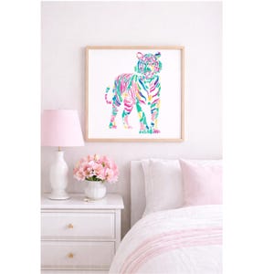 May include: A framed watercolour print of a tiger in vibrant colours, including pink, green, and yellow. The artwork is displayed above a white bedside table with a pink lamp and a bouquet of pink flowers. The bedroom setting has a white bedspread with pink accents.