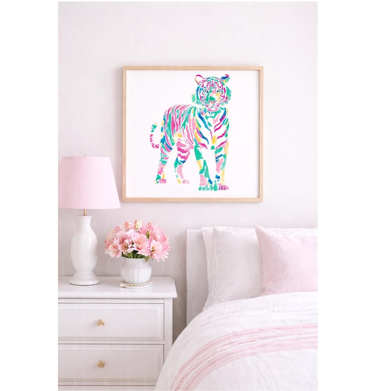 May include: A framed watercolour print of a tiger in vibrant colours, including pink, green, and yellow. The artwork is displayed above a white bedside table with a pink lamp and a bouquet of pink flowers. The bedroom setting has a white bedspread with pink accents.
