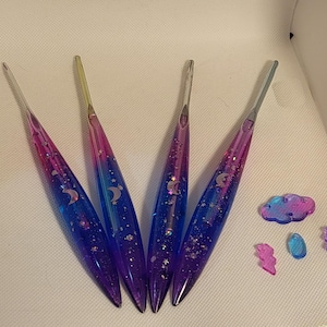 May include: Four crochet hooks with tapered, translucent handles featuring a gradient of purple, pink, and blue. The handles are embedded with silver glitter, star shapes, and crescent moon icons. Beside them are small cloud, raindrop, and lightning bolt charms.