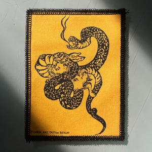 Könnte beinhalten: Ein rechteckiger Aufnäher mit gelbem Hintergrund und schwarzem, gesticktem Rand. Der Aufnäher zeigt eine schwarze Illustration einer Schlange, die sich um einen Tiger windet. Der Text "DARK ART TATTOO. BERLIN" steht unten.