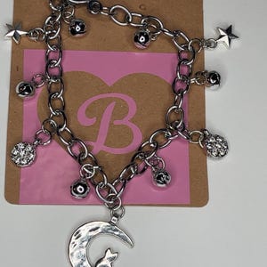 May include: Silver-toned bracelet with a toggle clasp, adorned with charms including stars, small bells, and a crescent moon with a cat silhouette. The bracelet is displayed on a pink heart-shaped background with a letter "B".