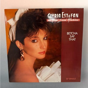 May include: Album cover for "Betcha Say That" by Gloria Estefan and Miami Sound Machine. The cover features a portrait of a woman with dark hair and red lipstick. The album title and artist name are printed in white and red.