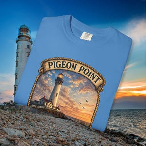 May include: A light blue t-shirt featuring a graphic of the Pigeon Point Lighthouse with a sunset backdrop. The lighthouse image is framed with a rope design and the words "Pigeon Point" in a banner. The shirt is displayed on a rocky surface.